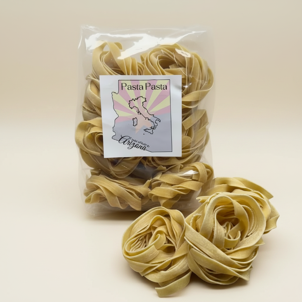 A clear plastic bag of flat pasta with a white label reading 'Pasta Pasta' and 'Made in Arizona,' with several loose rose-shaped pasta nests placed in front on a wooden surface against a white tiled background.