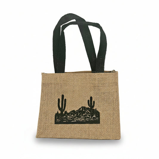 A natural jute tote bag with black fabric handles and a black printed desert landscape graphic featuring cacti and mountains, set against a plain off-white background.